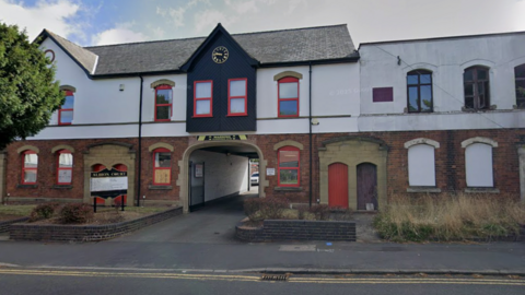 A part brick, part white rendered building, with red window frames. An access arch in the centre, with a clock is above it