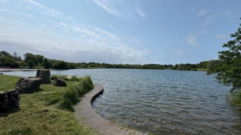 A general view of the Cosmeston Lakes Country Park. The lake is in the background and to the left of the picture there is a path, a grassy verge with some rocks on it.