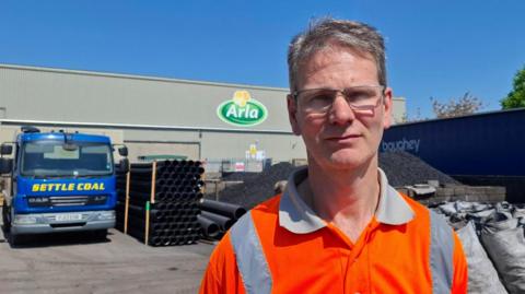 A man with short grey hair and glasses looks at the camera. He is wearing a hi-vis orange polo shirt. Behind him is a pile of coal, a lorry with the words "Settle Coal" on it, and a building with the Arla logo on the side.