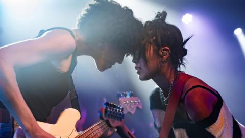 Two female guitar players touch heads on stage during a performance.