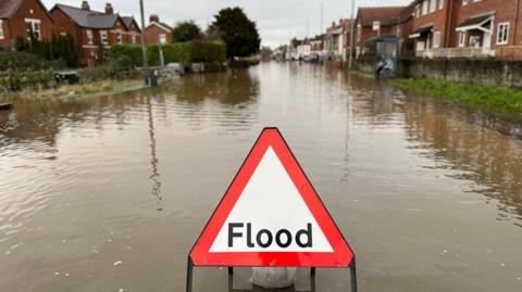 A triangle warning sign saying 'flood' in middle. The sign has been placed in flood water on a road in front of homes.