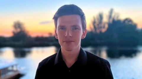 A white man with short black hair. He is looking at the camera and wearing a black shirt. He is standing in front of a lake. Behind the lake are black silhouettes of trees and a yellow and orange sunset