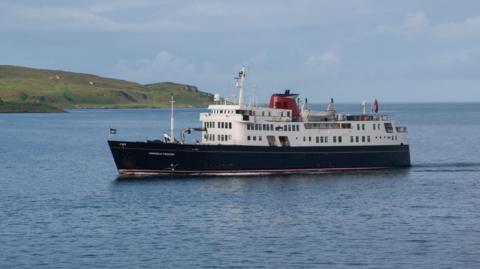 The small cruise ship is blue and white with a red funnel. It is moving across a calm sea. and there is a grassy headland in the distance.