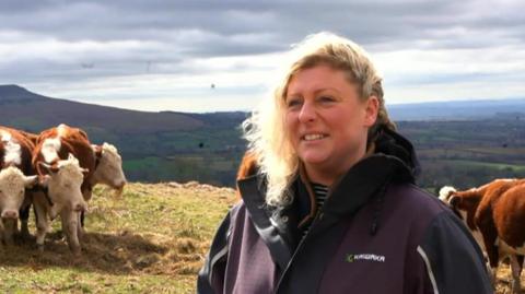 A mid shot of a woman standing in a hillside field next to cows. She has wavy blonde hair and is wearing a green and purple waterproof jacket. She is standing side-on to the camera. 