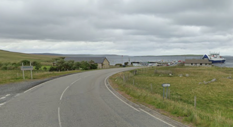 The road near Toft ferry terminal. The roads runs through an area of fields. There is a blue and white ferry at the terminal. It is a grey, cloudy day.