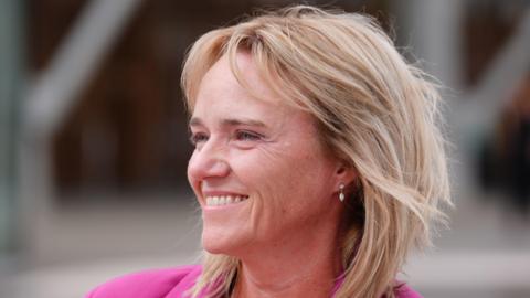 Sandie Peggie smiles and laughs while standing outside - she is wearing a pink jacket and has blonde hair