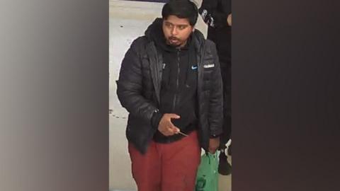 Missing man Yusef is seen on a CCTV camera walking and looking slightly to his right. He has thick dark smooth hair and a moustache and goatee beard. He is wearing a black hoodie and open black puffa style jacket, and red trousers. He is carrying a green carrier back and there is the elbow of another person to his left. 