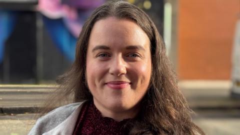 Áine Murray looks at the camera, smiling slightly. She has long, dark hair and is wearing a red patterned top and a light grey top.