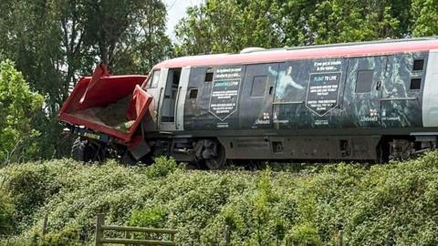 A train which has hit a red trailer. The trailer has been damaged by the train and is crumpled.