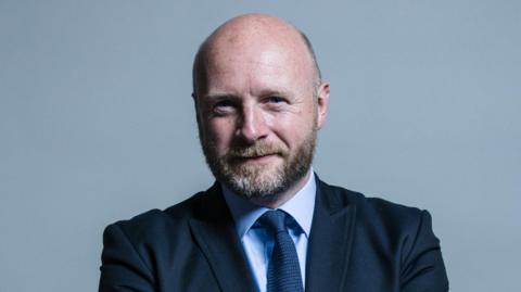 A portrait style photo of Liam Byrne, who is wearing a dark suit, pale blue shirt and navy tie, posed against a grey backdrop. He has a beard and balding head.