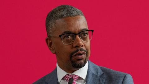 Vaughan Gething wearing a grey suit, white shirt and a red flowery tie. He is wearing glasses with black rims at the top and is standing in front of a red background.