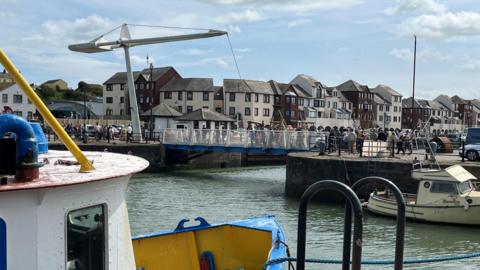 People cross the footbridge on Maryport's Elizabeth Dock. In the foreground of the image is a trawler berthed in the harbour. Town houses line the background.