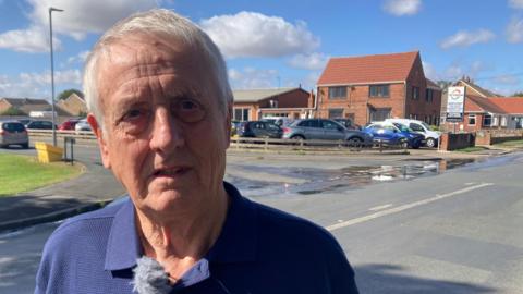 A man with short white hair and wearing a blue polo shirt stands in a village street in front of a spill of water, which covers the entrance to a junction. On the right is a green grass verge and in the background are red brick houses. Cars are parked on the street and in a small car park.