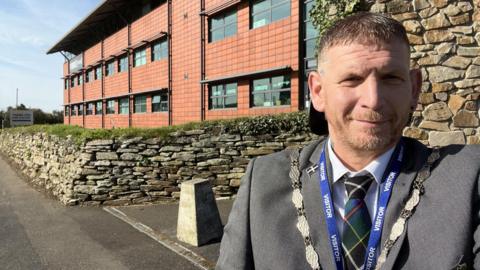 Mayor of Camborne James Ball stood in front of one of the schools wearing a grey suit and his mayoral chain. The school is a modern building clad in red.