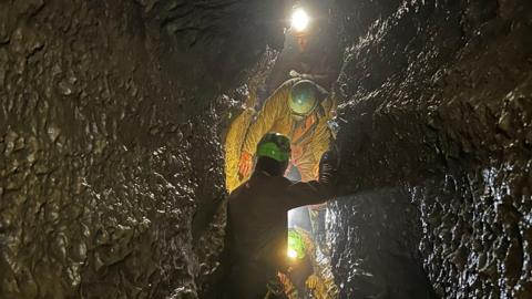 A narrow, rocky passageway within a cave. Two individuals are present, both wearing helmets fitted with headlamps that cast light on the dark, confined surroundings. The person in the foreground is carrying a white bag and appears to be carefully manoeuvring through the tight space. Further ahead, another person is visible, also making their way through the passage.
The cave walls are wet and glistening.