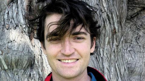 A close up image of Ross Ulbricht. He has dark hair which is being swept infront of his eyes, perhaps by the wind, and is standing infront of a tree trunk.