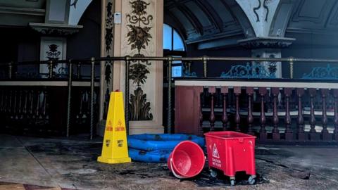 The inside of the Kursaal, a dimly lit atrium with deteriorated flooring. In the foreground is a yellow "caution" cone with two red buckets and a inflatable paddling pool being used to catch dripping water. 