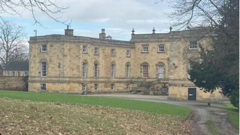 Historic three story stone building with lots of windows. There are trees to the right and grass in front with a path/road leading up to the building. 