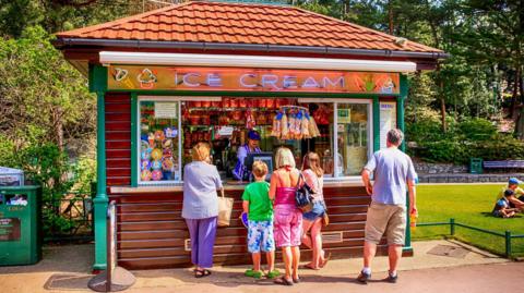 A lady and two children are at the counter opf an ice cream kiosk situated in parkland with a neon sign above that reads 'ice cream'. A man holding a drink stand to the right hand side and a lady with a shopping bag is also at the counter as a sales assistant takes orders.