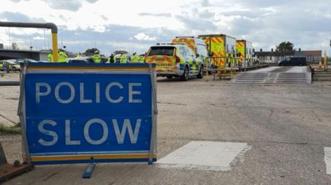 A Police Slow sign, in blue with white text, on a concrete hardstanding, with several police vehicles in the distance, and a weighbridge ramp on the right of the picture.
