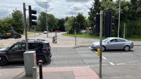 A gyratory system with multiple lanes. In the foreground, two cars drive through a set of traffic lights with cars in the background taking either the right or left exist off the gyratory with a mother using a crossing point in the middle ground.