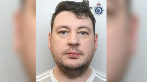 Thomas Hall faces the camera in a police mugshot. He has brown hair and facial stubble and is wearing a grey and white striped T-shirt.