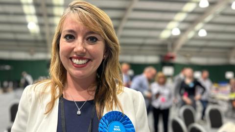 A smiling woman with long blonde hair wearing a white blazer, a navy top and a blue rosette standing in a busy hall.