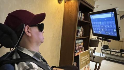 Bram Harrison in his wheelchair, in front of an eye-controlled computer screen with boxes which can be controlled with the gaze of his left eye.