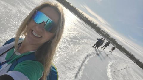 Annette Mawby, a woman wearing green glasses and standing on snow while taking a selfie - with more people seen in the background of her picture.