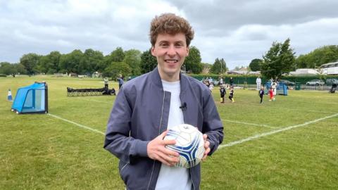 BBC's Jamie Moreland holds a football in front of a children's football pitch.