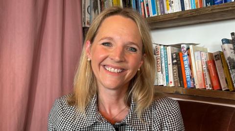 Kate Wright smiles at the camera. She is sat on a brown velvet chair, and there are bookshelves full of books behind her. She is wearing a short-sleeved gingham black and white button up shirt. 