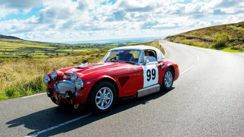 A red classic car with the number 99 painted on the side driving along a country road. 