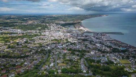 An aerial view of Ramsey, you can see a lot of houses along the curving coastline, with a pier that goes out into the sea.