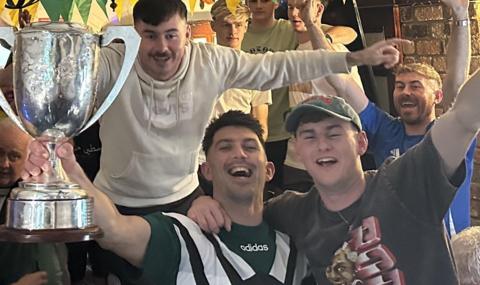 A packed scene inside a pub - in the foreground are two men, with beaming smiles. The one on the right has his left arm aloft and his right arm around the neck of the other man. The other man is holding aloft a trophy. In the background are several other men, who have their arms up in triumph. In the background we can see some bunting in the yellow and green, the colours of their club Dunloy. 
