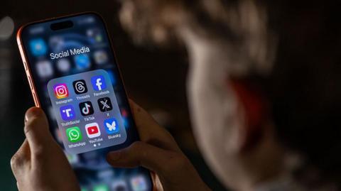 A child, who is out of focus, looks at a phone held in both hands. On the screen is a folder labelled 'social media' with app icons for Instagram, Threads, Facebook, TruthSocial, TikTok, X, WhatsApp, YouTube and Bluesky.