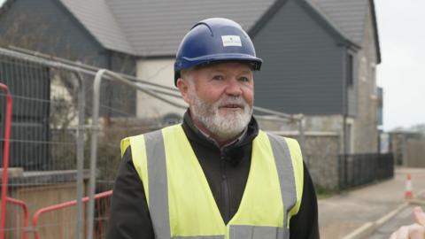 A man in a high viz jacket and a hard hat stands in front of a new housing development. He has a trimmed white beard.