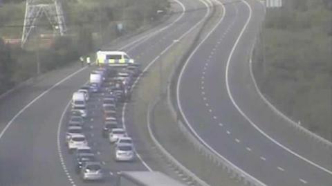 A traffic camera picture of the M49, showing traffic being held by police. A row of cars is shown queued up behind a police van parked sideways on the road. 