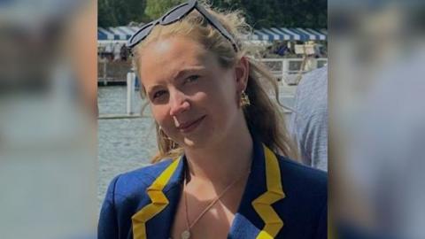 Rachael Debenham smiling at the camera. She is blonde haired, wearing a blue blazer with yellow trim, there is a river in the background. She has sunglasses on her head. 