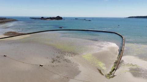 A drone shot looks down on a beach. A large, arcing concrete wall - which is supposed to hold water - starts on the sand and curves into and back out of sea. On the sand, a long yellow rope with several buoys lies along the sand on the entrance to the structure. The sea is turquoise under the blue sky, with a castle in the sea in the background.
