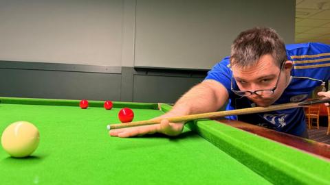 Dylan leaning low over a snooker table, aiming a cue stick at a white ball in the foreground, with three red balls positioned further behind on the green felt table. Dylan is wearing a blue sports t-shirt and black-framed specs low on his nose. He is in a brightly lit indoor venue.