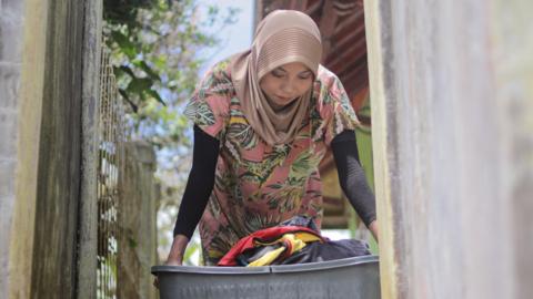 Young Asian Muslim woman wearing a hijab and casual daily home outfit holds a plastic laundry basket filled with clothes in the morning