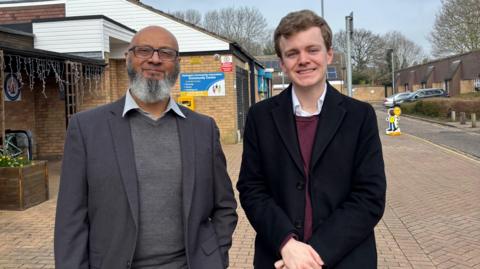 Ejaz, a man, wearing a grey blazer and grey cardigan, standing next to Sam Carling, a man in a black  coat, smiling for the camera.