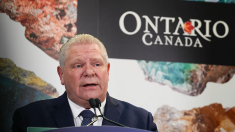 Doug Ford wears a dark blue suit and stands in front of a microphone. Behind him, there's a large sign that reads Ontario, Canada.