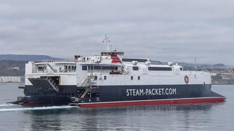 The Manannan sailing out of Douglas Harbour. It it a catamaran painted in white, red and black, and has STEAM-PACKET.COM on the side. 