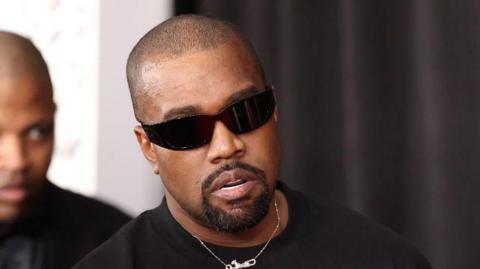 Kanye West is wearing black sunglasses and a black t-shirt. He has a shaved head and goatee, and his mouth is slightly open. There is a thin silver necklace with a silver pendant peeking over the t-shirt. He is walking somewhere and although the background is blurred, we can see someone walking behind him. There are a black curtain and a white wall in the background, too.