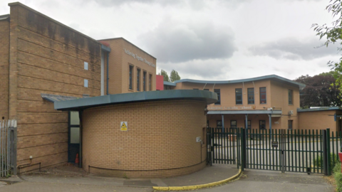 The exterior of St Edmund Campion Catholic School in Erdington. A small round brick building is next to a secure green gate, leading into a playground in front of the main school building.