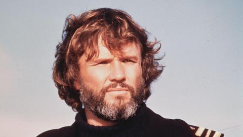 Kris Kristofferson staring off into the distance, squinting a little like the sun is in his eyes. He has shaggy brown hair, a greying beard and the sky is light blue behind him.