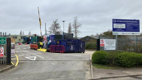 The entrance to a recycling centre, with a large sign to the right, blue containers at the entrance, a barrier up in the distance, and a green cabin on the left. There are several trees in the distance. There are lots of signs and a lamp post in the middle.