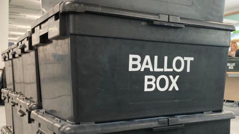 A number of black plastic ballot boxes stacked on top of one another. The words 'BALLOT BOX' are printed in white on the side. 