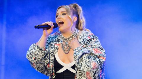 Ella Henderson sings on stage with a blue background. She has blonde hair in a ponytail and several silver chains around her neck. She is wearing a large white-coloured coat covered in red and black writing.